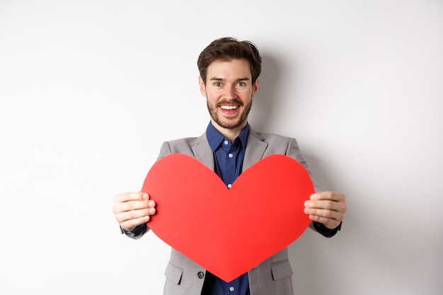 Jovem bonito, desejando feliz dia dos namorados, dando um grande sinal de coração vermelho e sorrindo, fazer surpresa para o amante, em pé no terno sobre fundo branco.