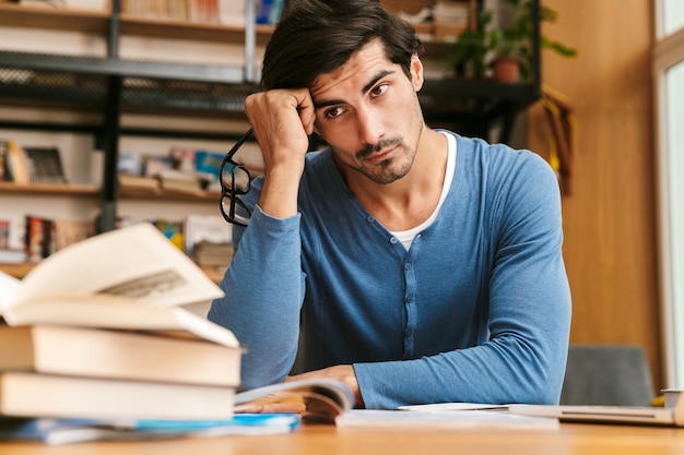 Jovem bonito e cansado sentado na mesa da biblioteca, trabalhando / estudando