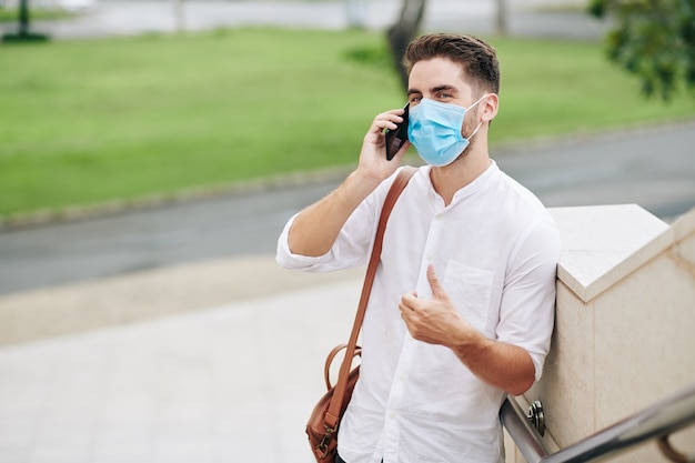 Jovem bonito e positivo numa máscara médica falando ao telefone com um colega ou amigo