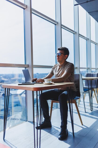 Jovem bonito e sorridente freelancer inteligente de óculos trabalha em um laptop enquanto passa o tempo em um café Um estudante positivo se senta em uma mesa de madeira em um café Homem feliz sentado enquanto trabalha dentro de casa