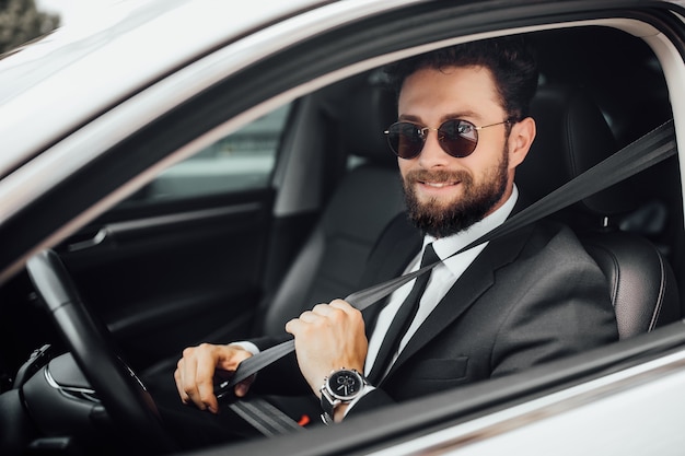 Jovem bonito e sorridente motorista barbudo com terno completo e cinto de segurança ao dirigir um carro