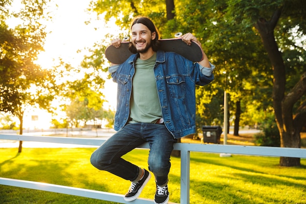 Foto jovem bonito e sorridente passando um tempo no parque, segurando um skate, sentado em um trilho