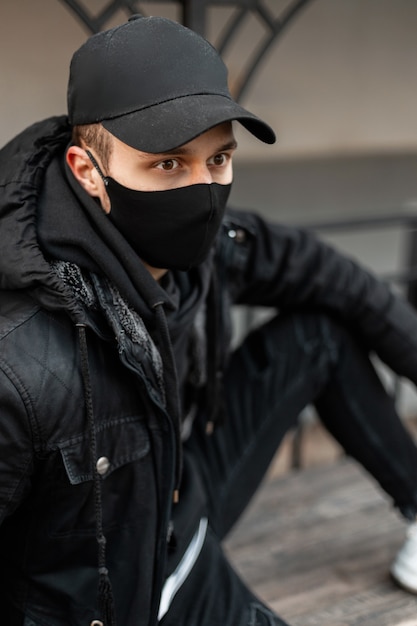 Jovem bonito elegante com máscara protetora preta na jaqueta preta da moda e moletom com capuz sentado na rua