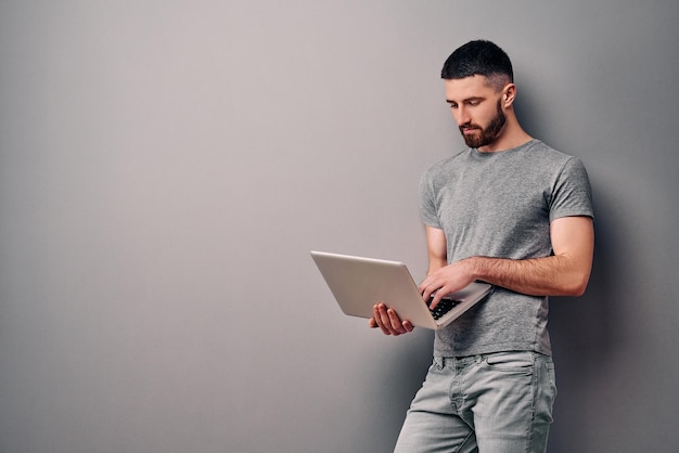 jovem bonito elegante em camiseta cinza em pé contra anúncios de espaço de cópia de parede cinza segurando laptop