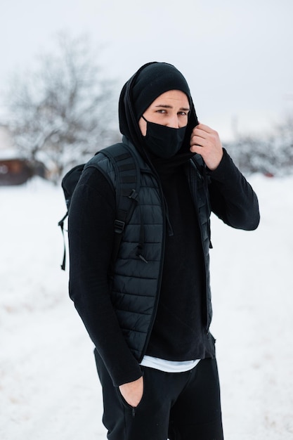 Jovem bonito elegante modelo masculino com máscara protetora preta em colete de moda preto, capuz, chapéu e mochila preta elegante viaja em dia de inverno nevado. Conceito de coronavírus e estilo