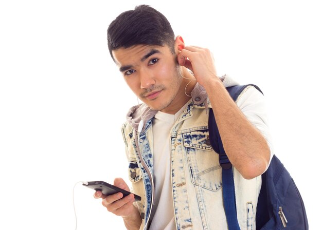 Jovem bonito em camiseta e jaqueta jeans com mochila azul usando smartphone e fones de ouvido