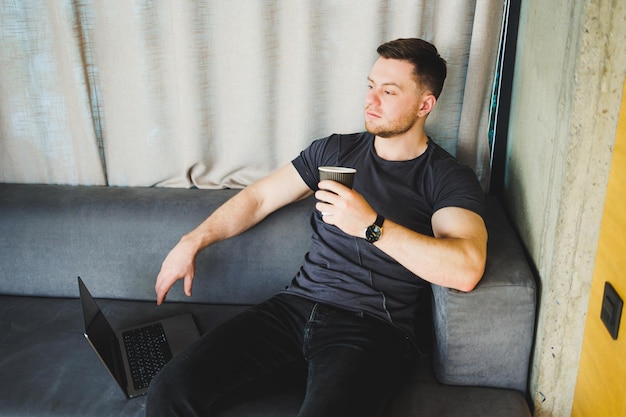 Jovem bonito em camiseta sentado no sofá e tomando café trabalhando remotamente durante a inicialização como freelancer olhando para laptop e sorrindo