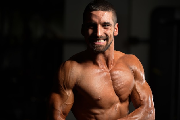 Jovem bonito em pé forte na academia e flexionando os músculos Muscular Atlético Fisiculturista Modelo de fitness posando após exercícios