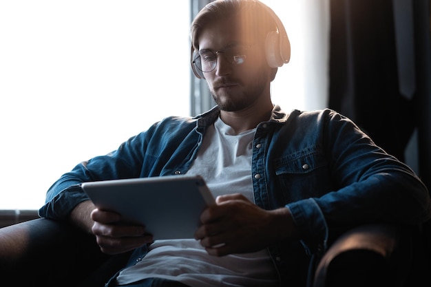 Jovem bonito em roupas casuais usando um tablet digital e fones de ouvido e sorrindo enquanto está sentado na cadeira