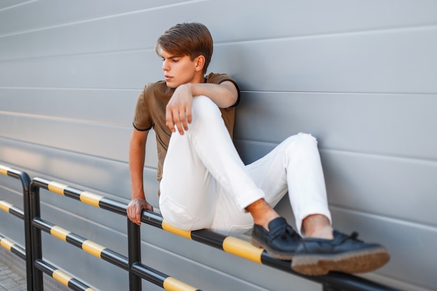 Jovem bonito em roupas da moda elegantes sentado perto de uma parede de metal