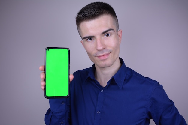 Jovem bonito em uma camisa azul apresenta smartphone com tela verde usando chroma key