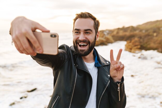 Jovem bonito em uma jaqueta de couro tirando uma foto de selfie no smartphone enquanto caminhava à beira-mar