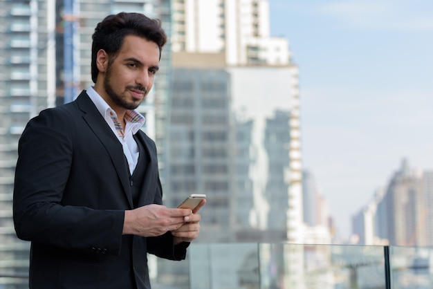 Jovem bonito empresário indiano segurando um telefone celular enquanto pensa em um telhado em Bangkok