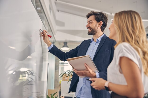 Jovem bonito escrevendo na lousa e sorrindo ao lado de uma colega de trabalho