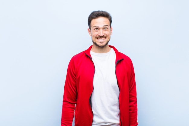 Jovem bonito esportes homem ou monitor olhando feliz e pateta com um sorriso largo, divertido e maluco e olhos bem abertos na parede plana