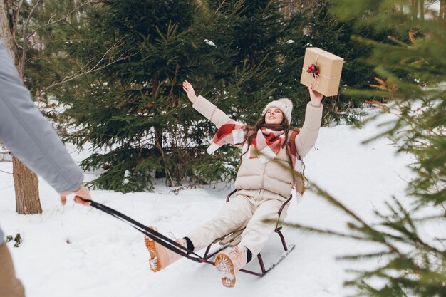 Jovem bonito está se divertindo em um trenó com a namorada com presentes em uma floresta de coníferas de inverno. Um parque com árvores de Natal ao fundo. Espírito de natal. Tingimento.