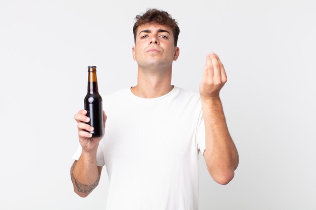 Jovem bonito fazendo um gesto de capice ou dinheiro, dizendo para você pagar e segurando uma garrafa de cerveja