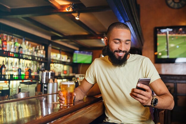 Jovem bonito feliz bebendo cerveja em bar e usando smartphone