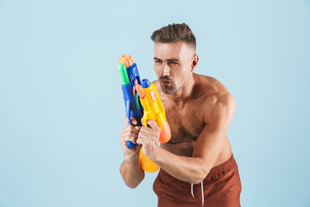 Jovem bonito feliz sem camisa usando shorts de praia em pé sobre o azul, brincando com pistolas de água