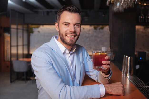 Jovem bonito feliz sorrindo para a câmera, torcendo com seu copo de cocktail