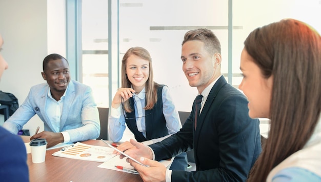 Jovem bonito gesticulando e discutindo algo enquanto seus colegas de trabalho o escutam sentado na mesa do escritório