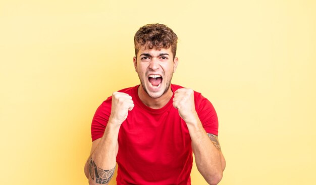 Jovem bonito gritando agressivamente com olhar irritado, frustrado e raivoso e punhos cerrados, sentindo-se furioso