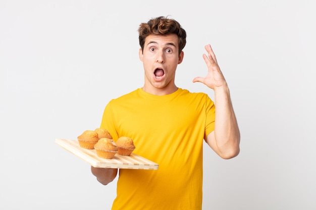 Jovem bonito gritando com as mãos para cima segurando uma bandeja de muffins