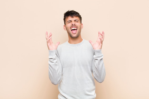 Jovem bonito gritando furiosamente, sentindo-se estressado e irritado com as mãos no ar dizendo por que eu na parede quente