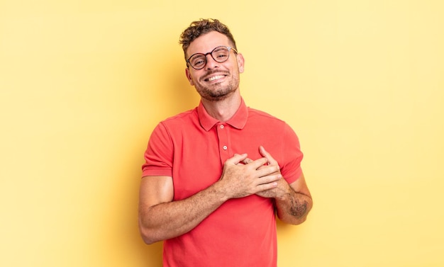 Jovem bonito hispânico se sentindo romântico, feliz e apaixonado, sorrindo alegremente e segurando o coração de mãos dadas