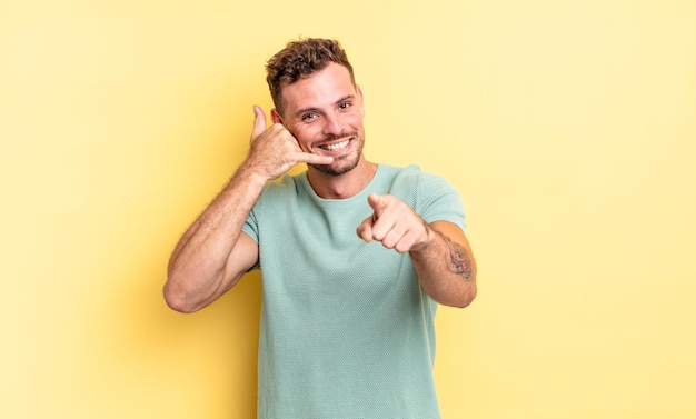 Jovem bonito hispânico sorrindo alegremente e apontando para a câmera enquanto faz um gesto depois de ligar para você, falando no telefone