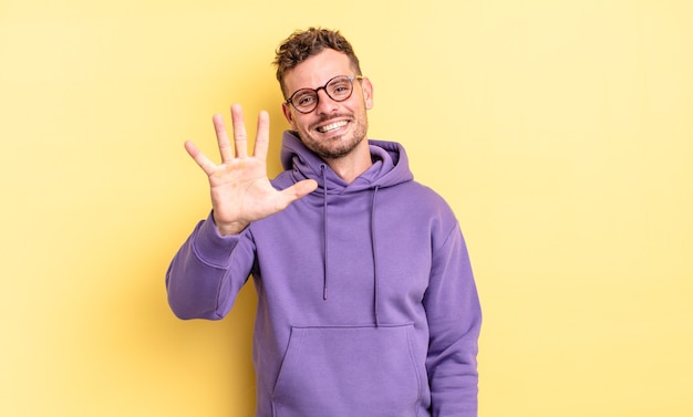 jovem bonito hispânico sorrindo e parecendo amigável, mostrando o número cinco ou quinto com a mão para a frente, em contagem regressiva