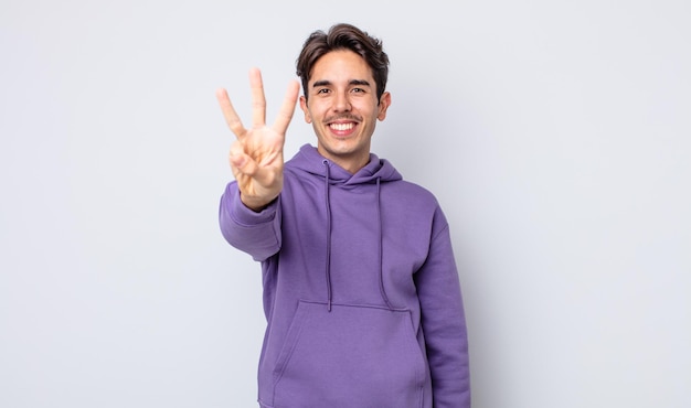 Jovem bonito hispânico sorrindo e parecendo amigável, mostrando o número três ou terceiro com a mão para a frente, em contagem regressiva