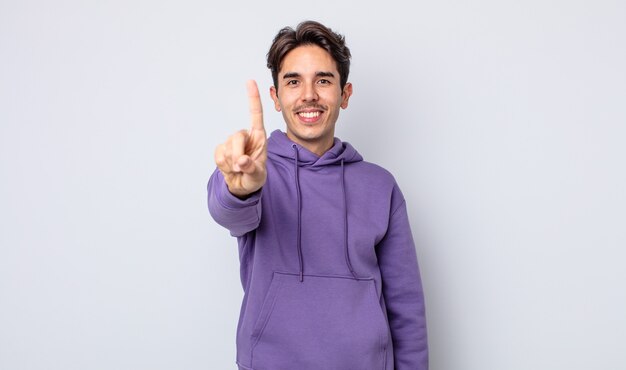 Jovem bonito hispânico sorrindo e parecendo amigável, mostrando o número um ou o primeiro com a mão para a frente, em contagem regressiva