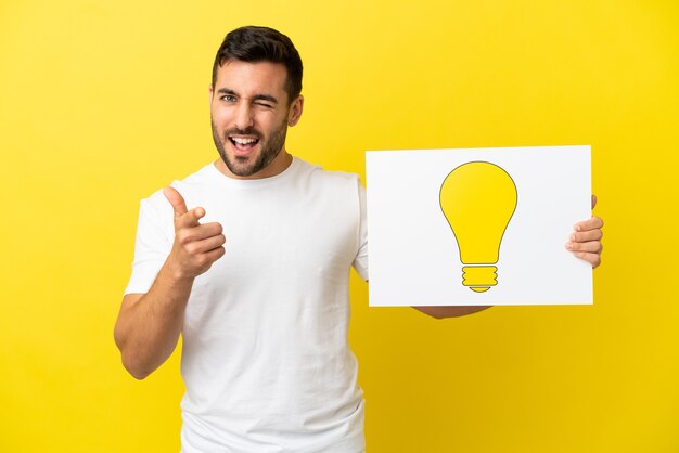 Jovem bonito homem caucasiano isolado em um fundo amarelo segurando um cartaz com o ícone de lâmpada e apontando para a frente