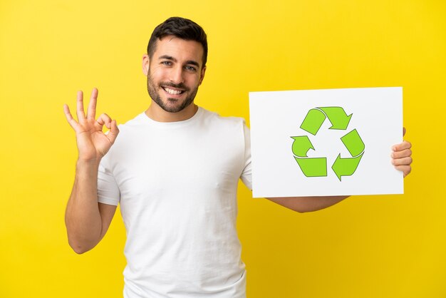 Jovem bonito homem caucasiano isolado em um fundo amarelo segurando um cartaz com o ícone de reciclagem e comemorando uma vitória