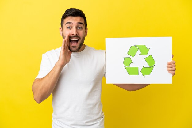 Jovem bonito homem caucasiano isolado em um fundo amarelo segurando um cartaz com o ícone de reciclagem e gritando