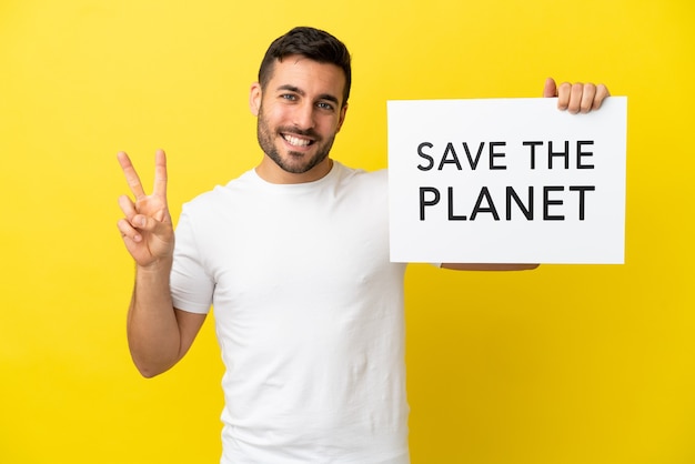 Jovem bonito homem caucasiano isolado em um fundo amarelo segurando um cartaz com o texto Salve o planeta e comemorando uma vitória