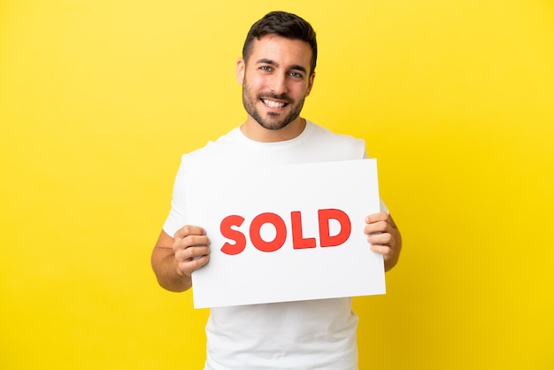 Jovem bonito homem caucasiano isolado em um fundo amarelo segurando um cartaz com o texto VENDIDO com uma expressão feliz