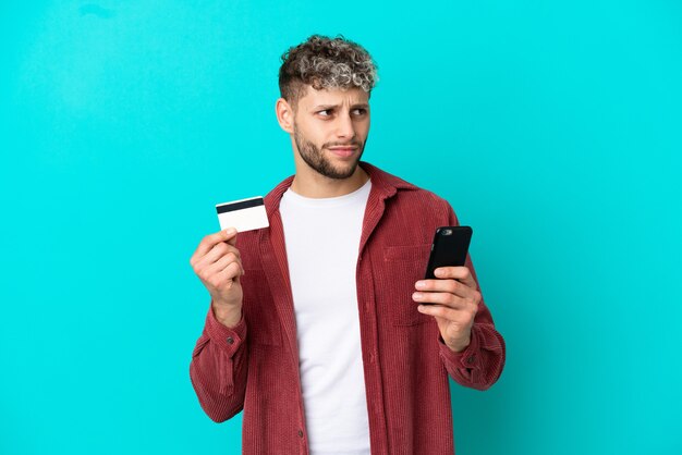 Jovem bonito homem caucasiano isolado em um fundo azul, comprando com o celular com um cartão de crédito enquanto pensa