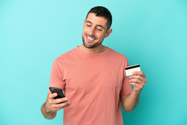 Jovem bonito homem caucasiano isolado em um fundo azul, comprando com o celular com um cartão de crédito