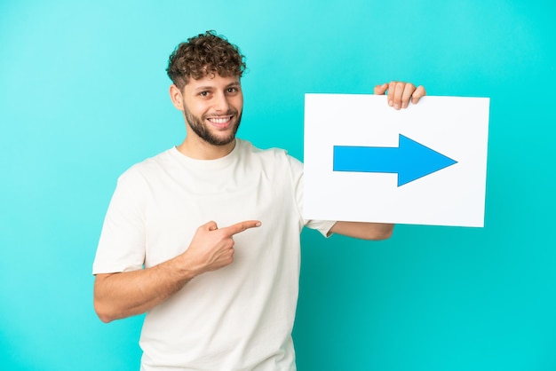 Jovem bonito homem caucasiano isolado em um fundo azul segurando um cartaz com o símbolo de uma seta e apontando-o