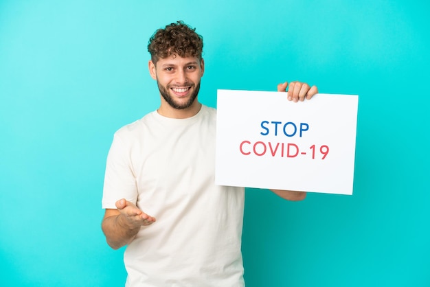 Jovem bonito homem caucasiano isolado em um fundo azul segurando um cartaz com o texto Pare Covid 19 fazendo um acordo