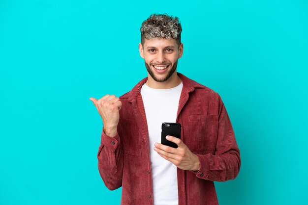 Jovem bonito homem caucasiano isolado em um fundo azul usando telefone celular e apontando para a lateral