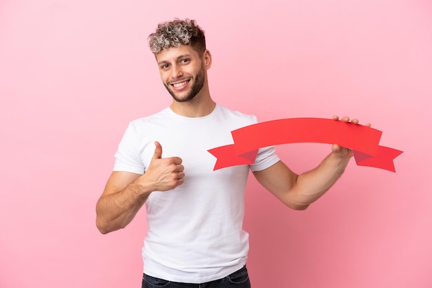 Jovem bonito homem caucasiano isolado em um fundo rosa segurando um cartaz vazio com o polegar para cima