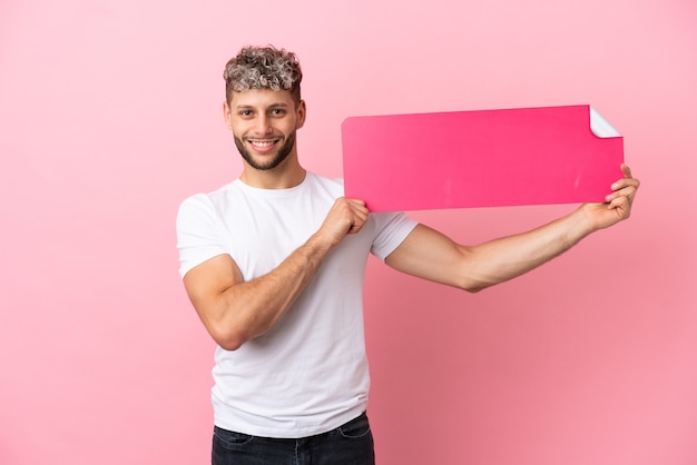 Jovem bonito homem caucasiano isolado em um fundo rosa segurando um cartaz vazio