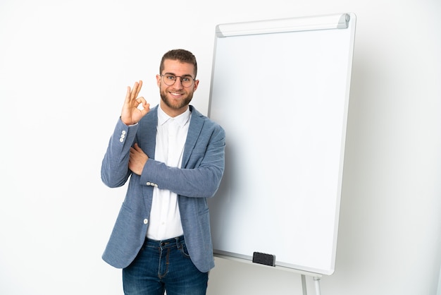 Jovem bonito homem caucasiano isolado no fundo branco fazendo uma apresentação no quadro branco e mostrando um sinal de ok com os dedos