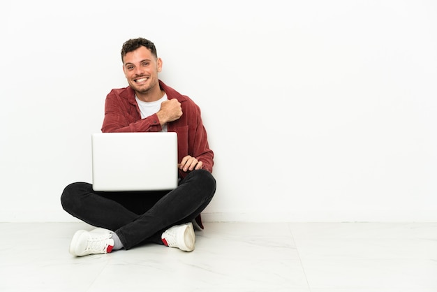 Jovem bonito homem caucasiano sentado no chão com laptop comemorando uma vitória