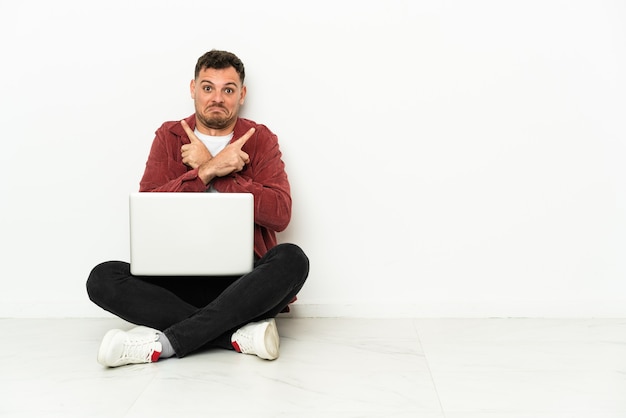 Jovem bonito homem caucasiano sentado no chão com o laptop apontando para as laterais, tendo dúvidas