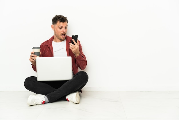 Jovem bonito homem caucasiano sentado no chão com um laptop segurando café