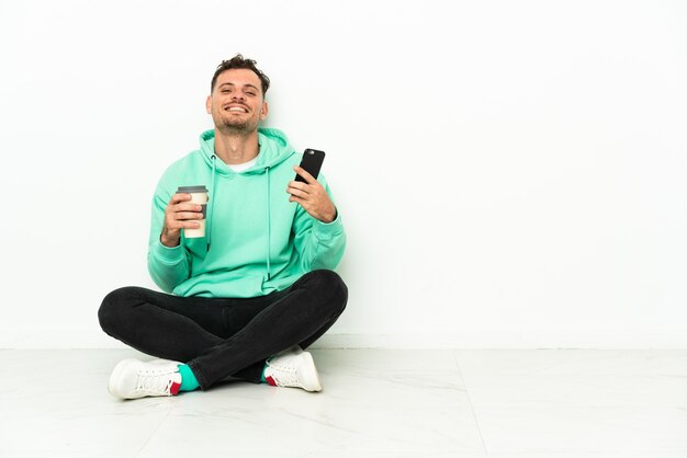 Jovem bonito homem caucasiano sentado no chão segurando um café para levar e um celular